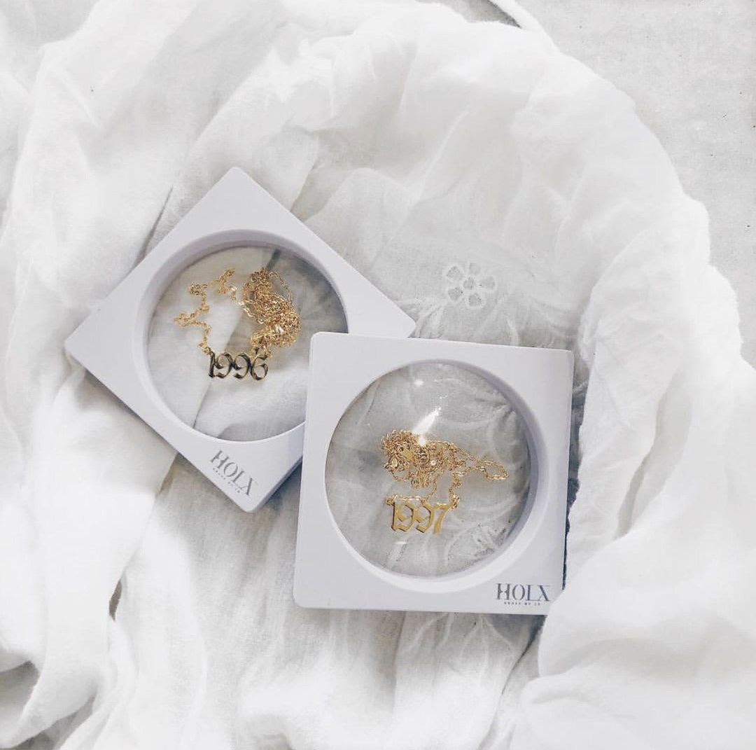 Two white jewelry boxes with gold necklaces inside, placed on a white fabric background.