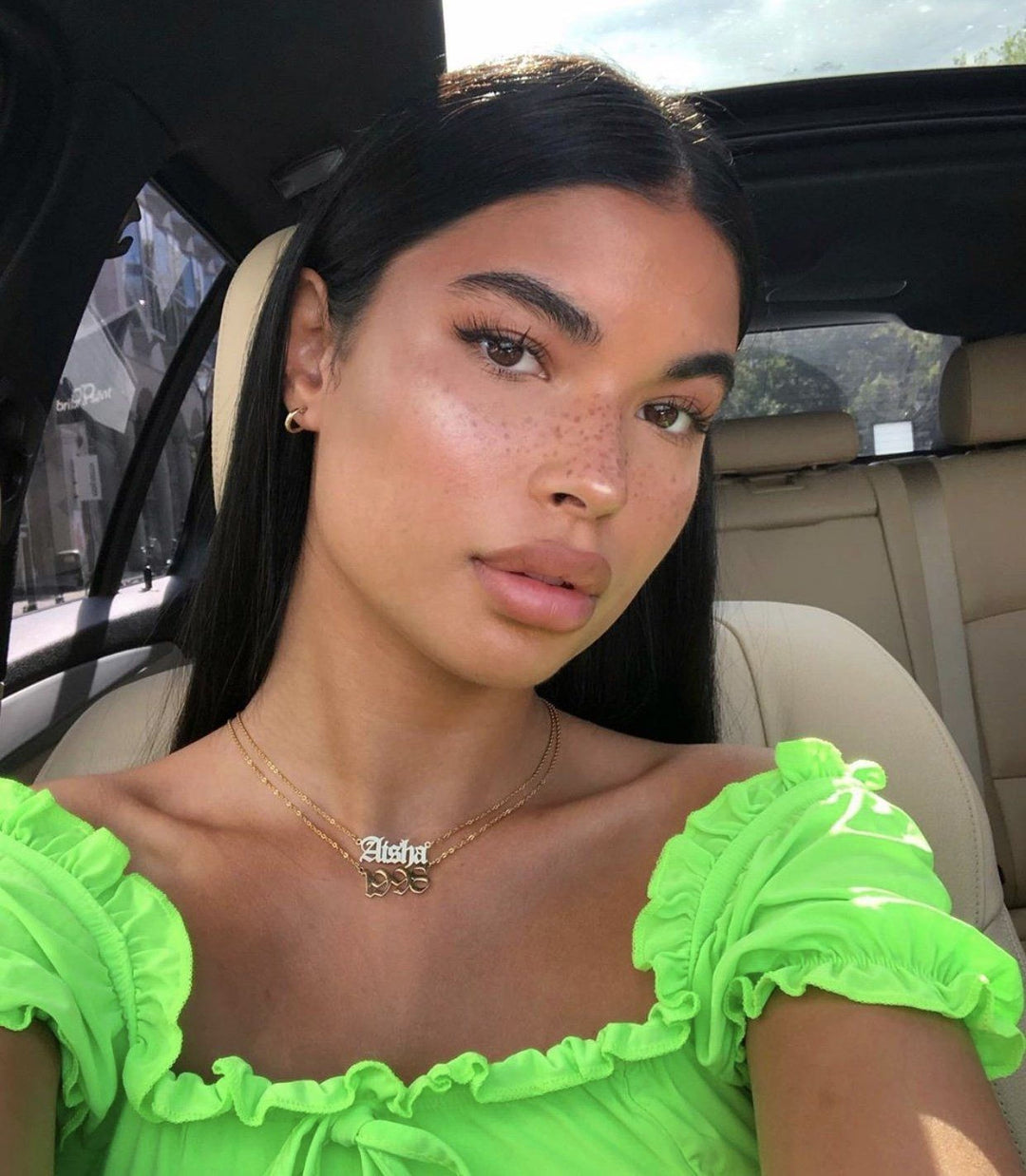 A young woman with long dark hair wearing a bright green off-the-shoulder top and a necklace with a pendant. She is sitting in the back of a car, looking directly at the camera.