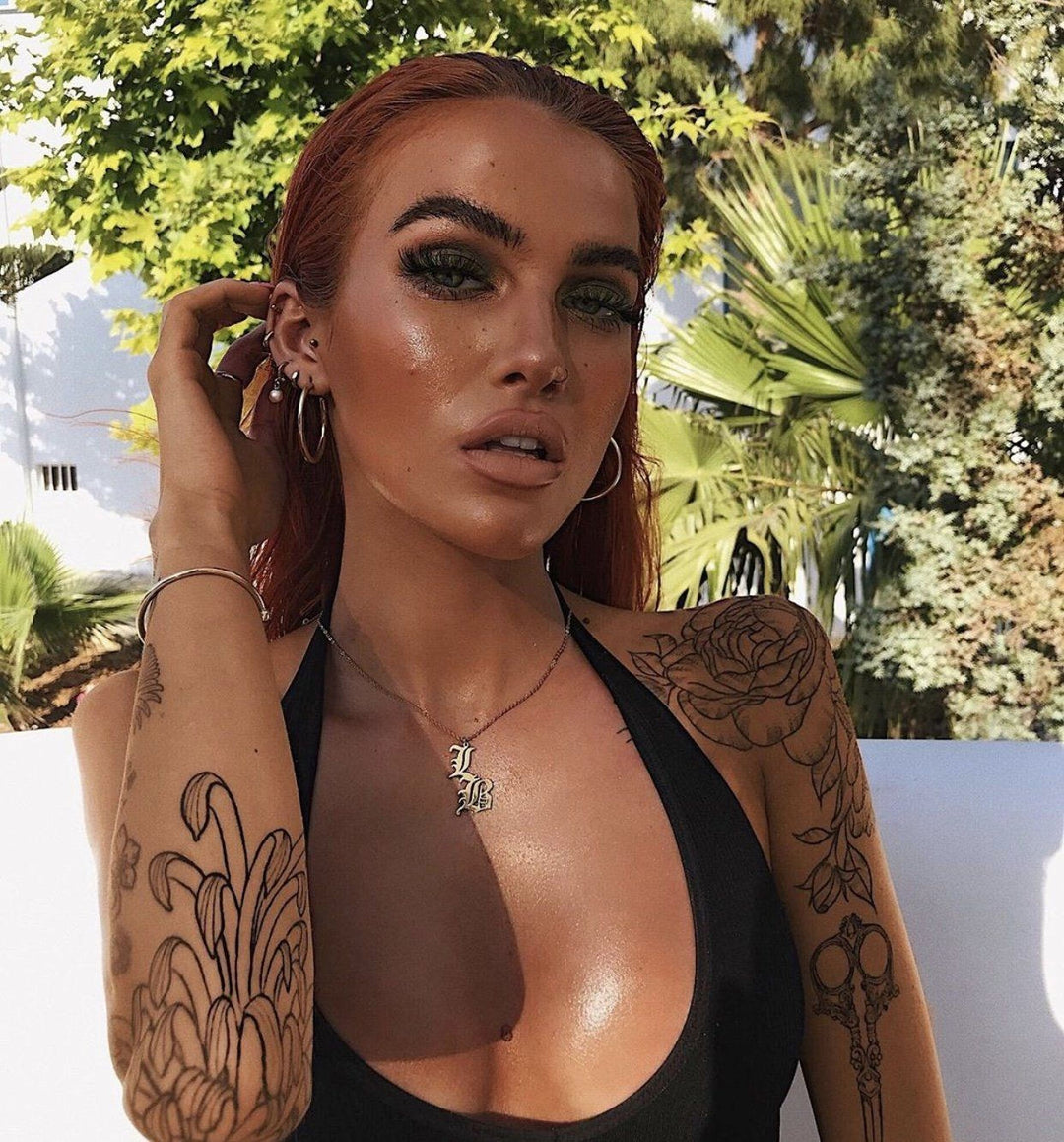 A woman with red hair and tattoos poses outdoors, wearing a black top and a necklace.
