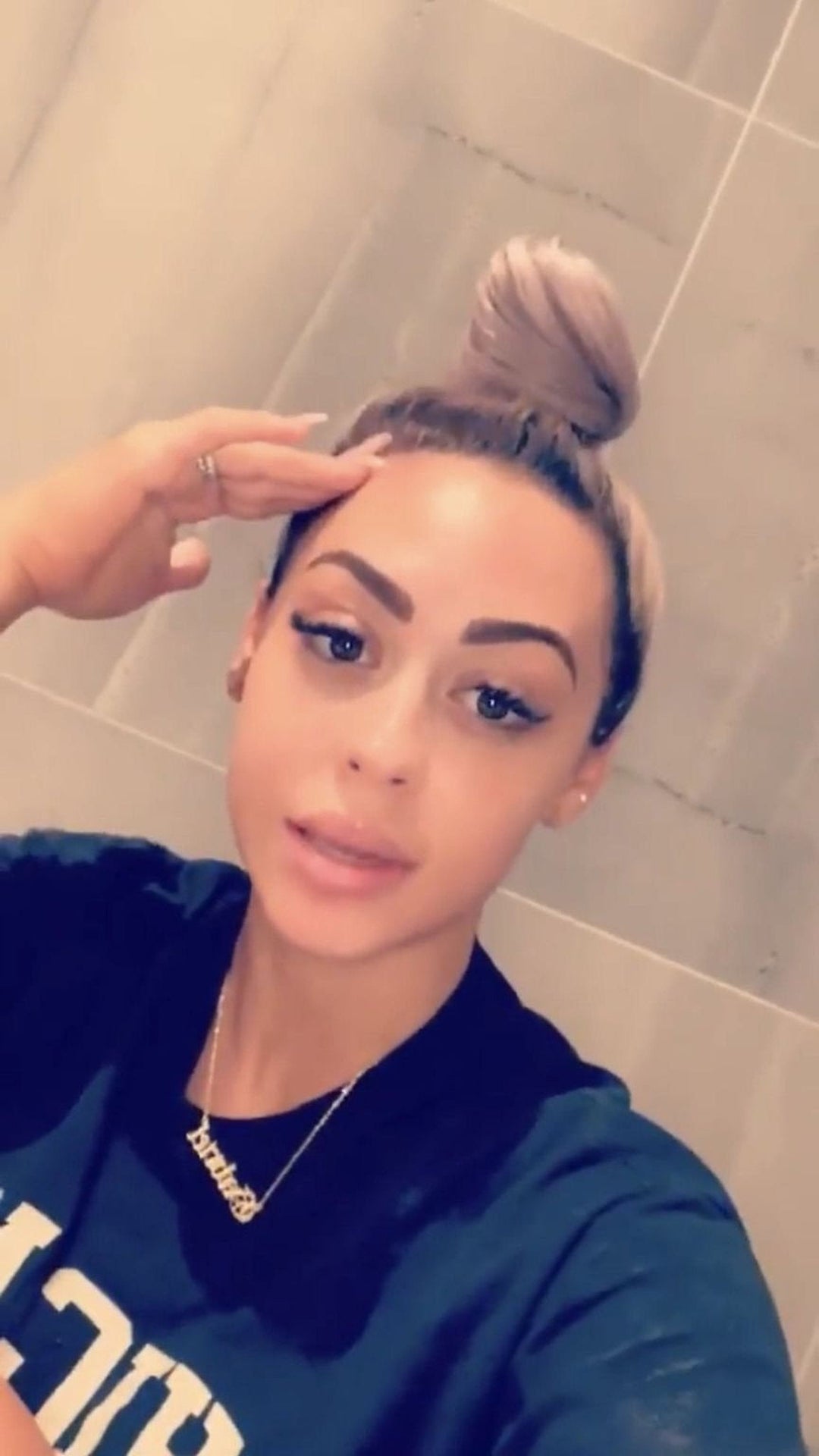 A woman with blonde hair is wearing a black shirt and has her hand on her forehead.