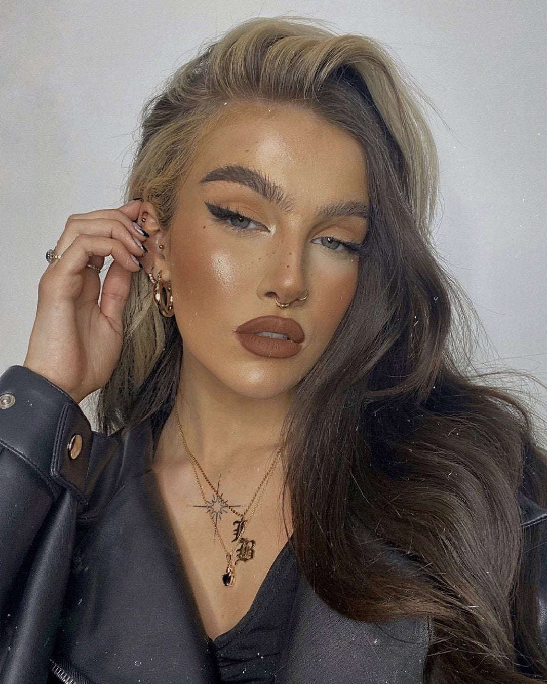 A woman with long, wavy brown hair and bold makeup poses for a portrait. She is wearing a black leather jacket and has a necklace with a star pendant.