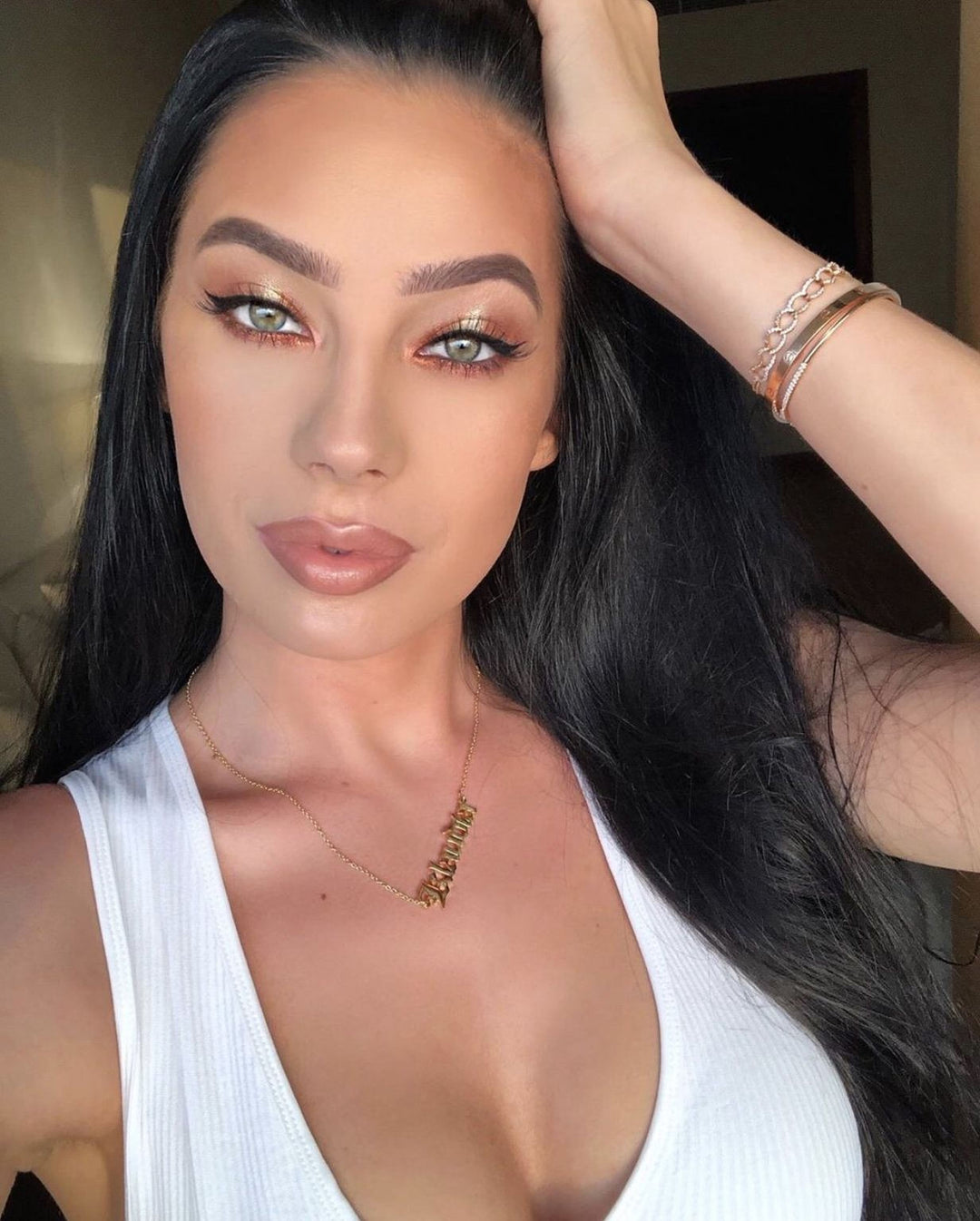 A woman with long dark hair wearing a white top and gold jewelry poses for a selfie.