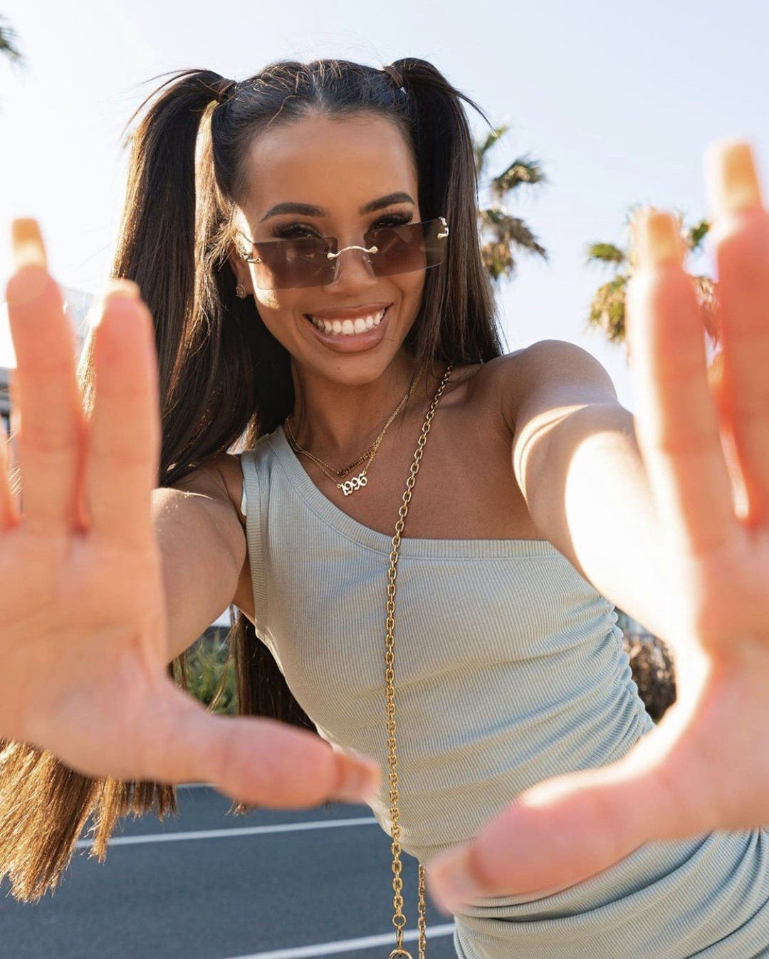 A woman with long brown hair and sunglasses is smiling and making a hand gesture, with her fingers spread wide.