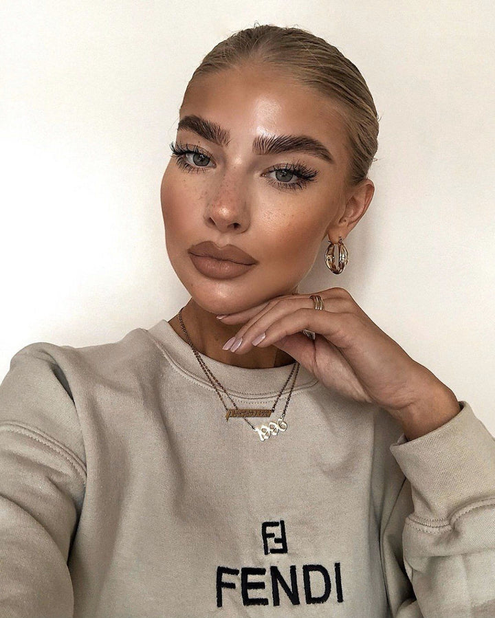 A woman with long blonde hair and makeup posing for a selfie. She is wearing a gray sweatshirt with the Fendi logo and has a hand resting on her chin.