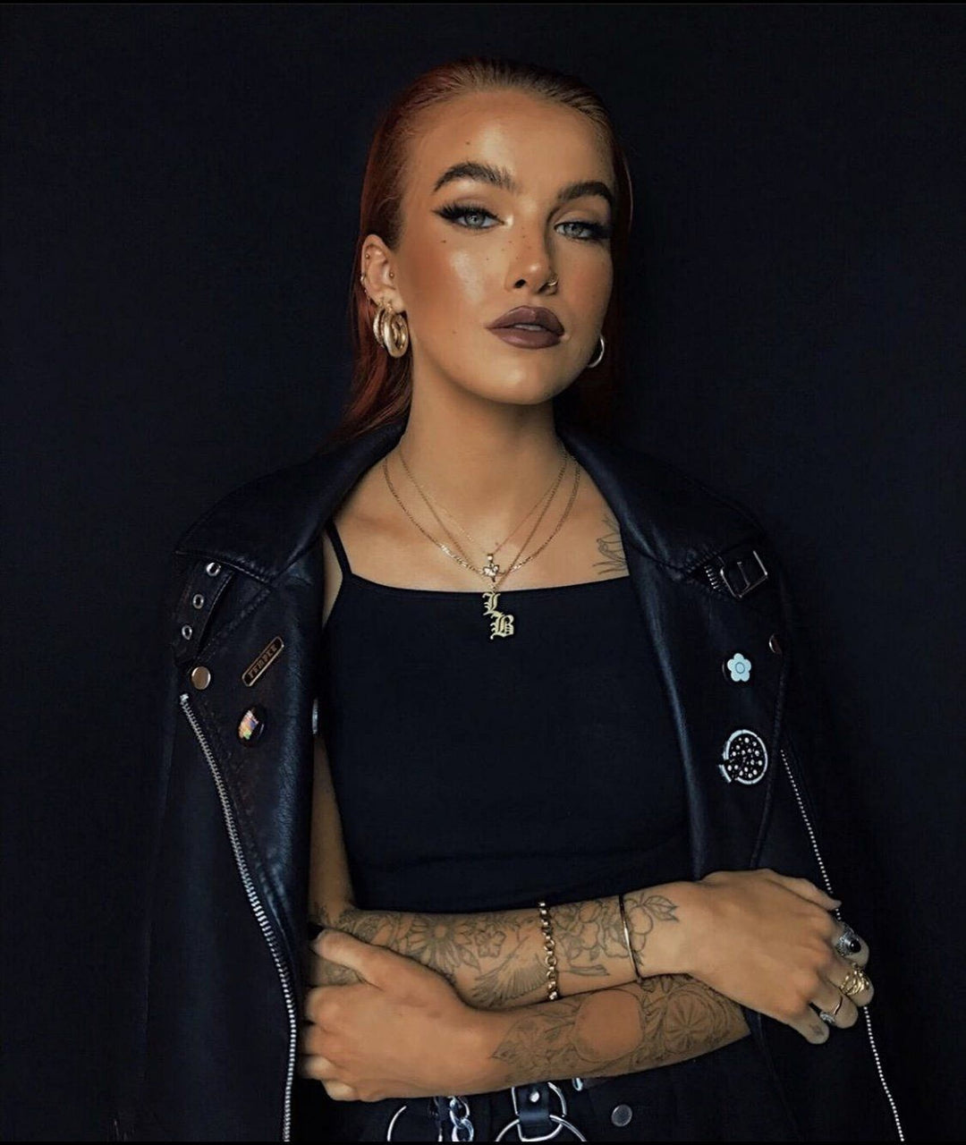 A woman with long red hair wearing a black leather jacket and a black top with a pendant necklace stands against a dark background.