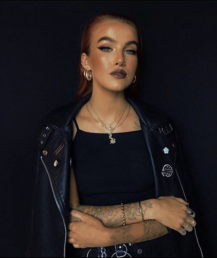 A woman with long red hair wearing a black leather jacket and a black top with a pendant necklace stands against a dark background.
