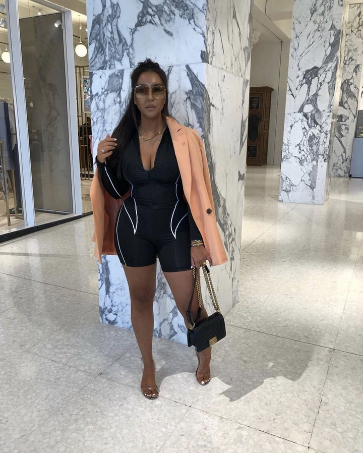 A woman wearing a black and white outfit, including a black jumpsuit and a peach jacket, stands in a marble-tiled room with a marble pillar. She is holding a black purse and wearing sunglasses.