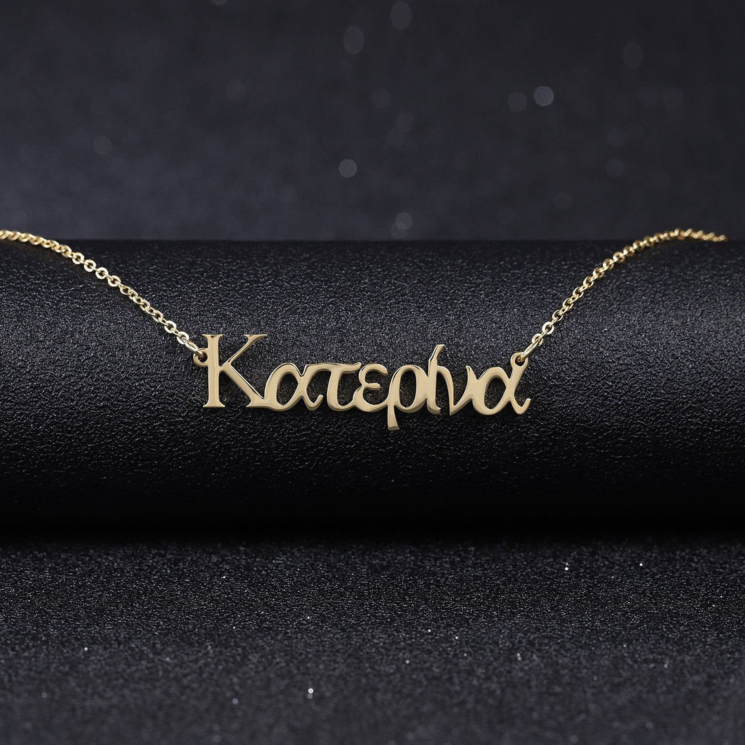 A gold-colored necklace with the word "Katerina" engraved on it, displayed on a black surface.