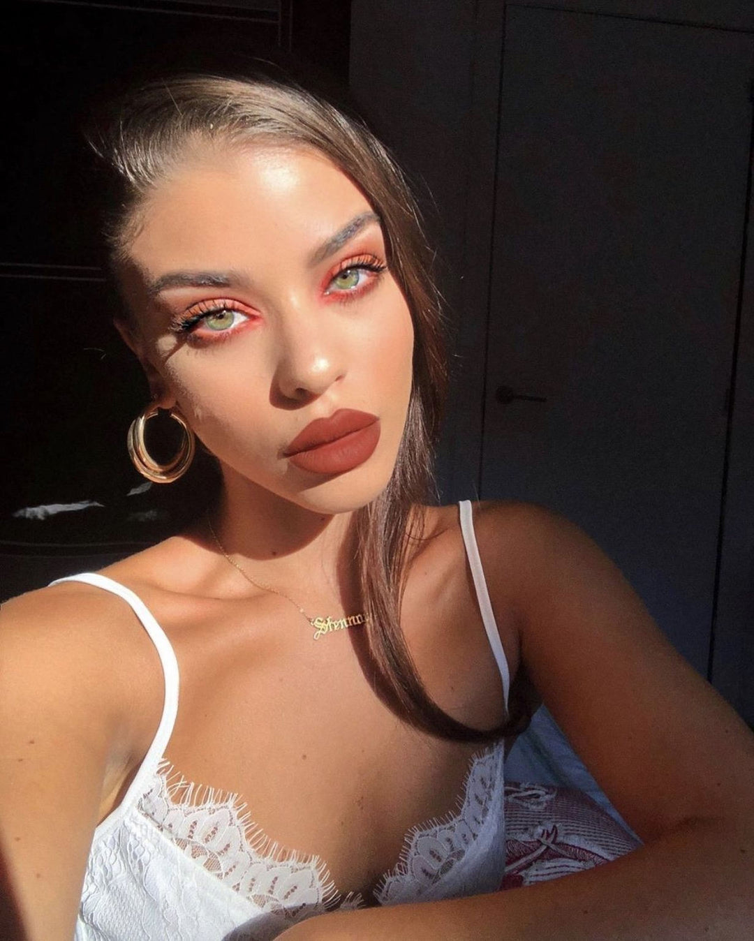A woman with long brown hair and striking red eyes wearing a white lace bra and large hoop earrings, posing for a selfie.