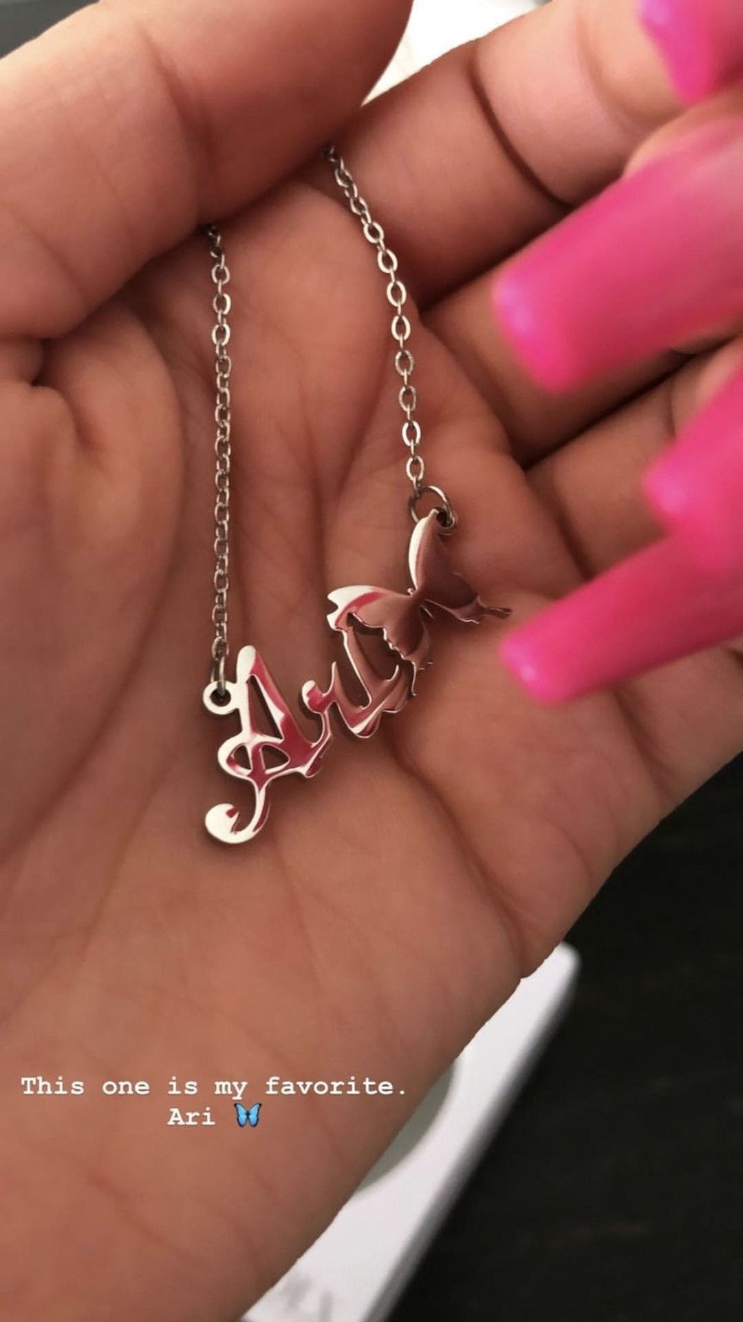 A hand holding a silver necklace with a butterfly pendant, and a caption below the image that reads "This one is my favorite Ari".