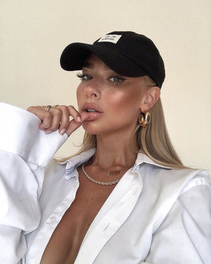 A woman wearing a black baseball cap, a white collared shirt, and gold hoop earrings poses for a portrait against a plain background.