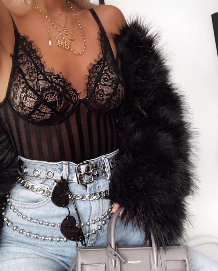 A woman wearing a black lace bra top, a black fur coat, and a chain belt, holding a gray handbag.