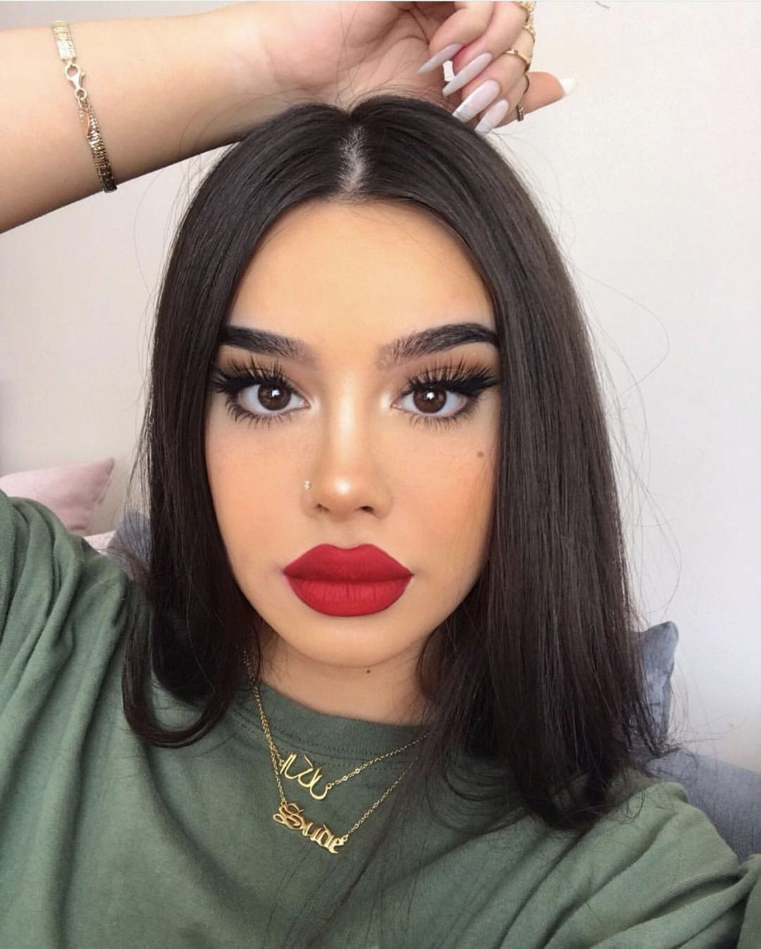 A woman with long dark hair and red lipstick is wearing a green shirt and has her hand on her head. She is also wearing a necklace with a pendant.