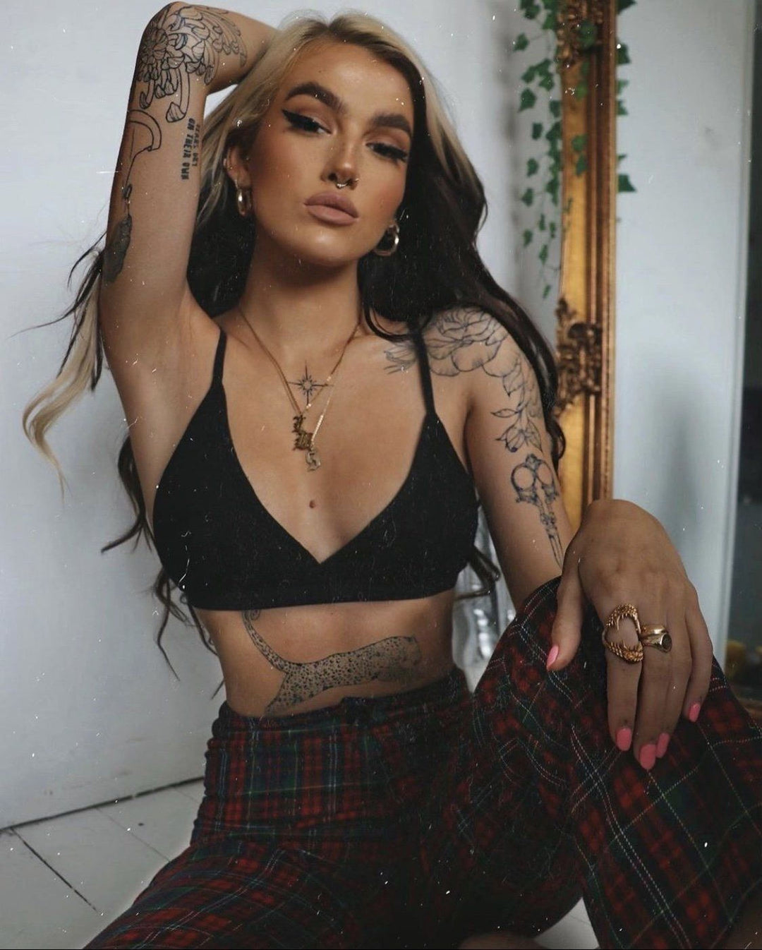A young woman with tattoos on her arms and legs is sitting on the floor, wearing a black crop top and plaid pants. She has long, dark hair and is looking directly at the camera.