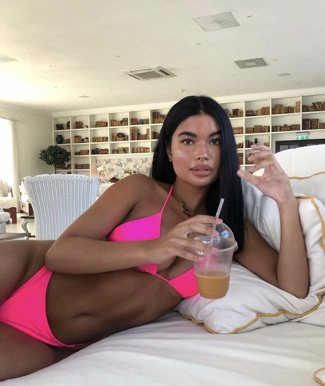 A woman in a pink bikini is sitting on a bed, holding a drink in her hand.