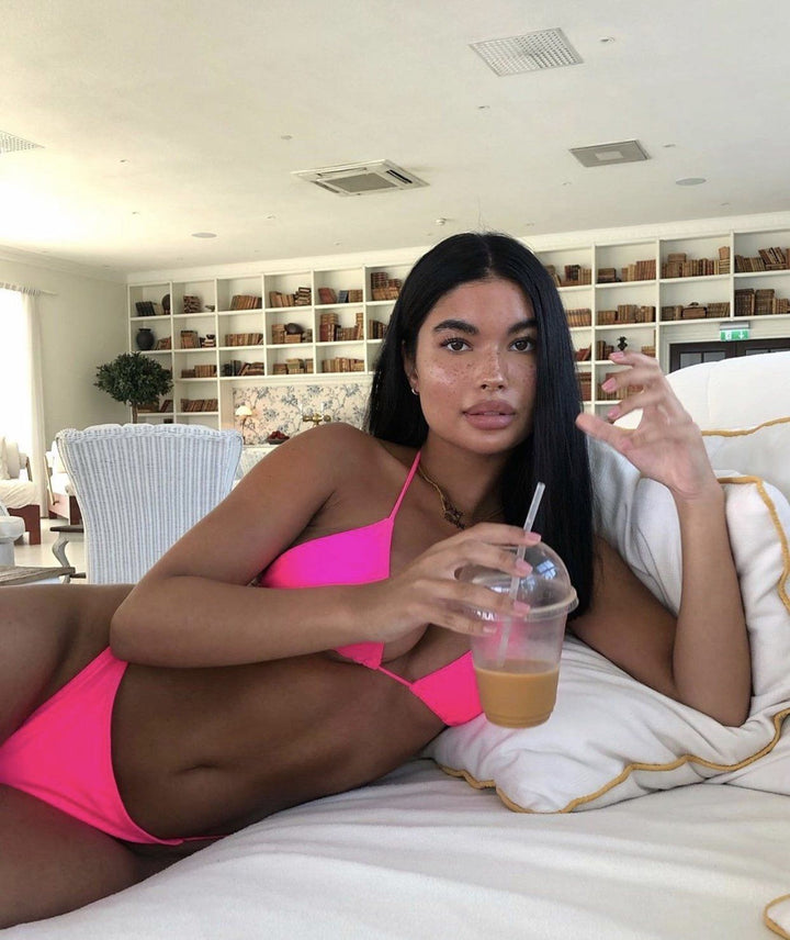 A woman in a pink bikini is sitting on a bed, holding a drink in her hand.