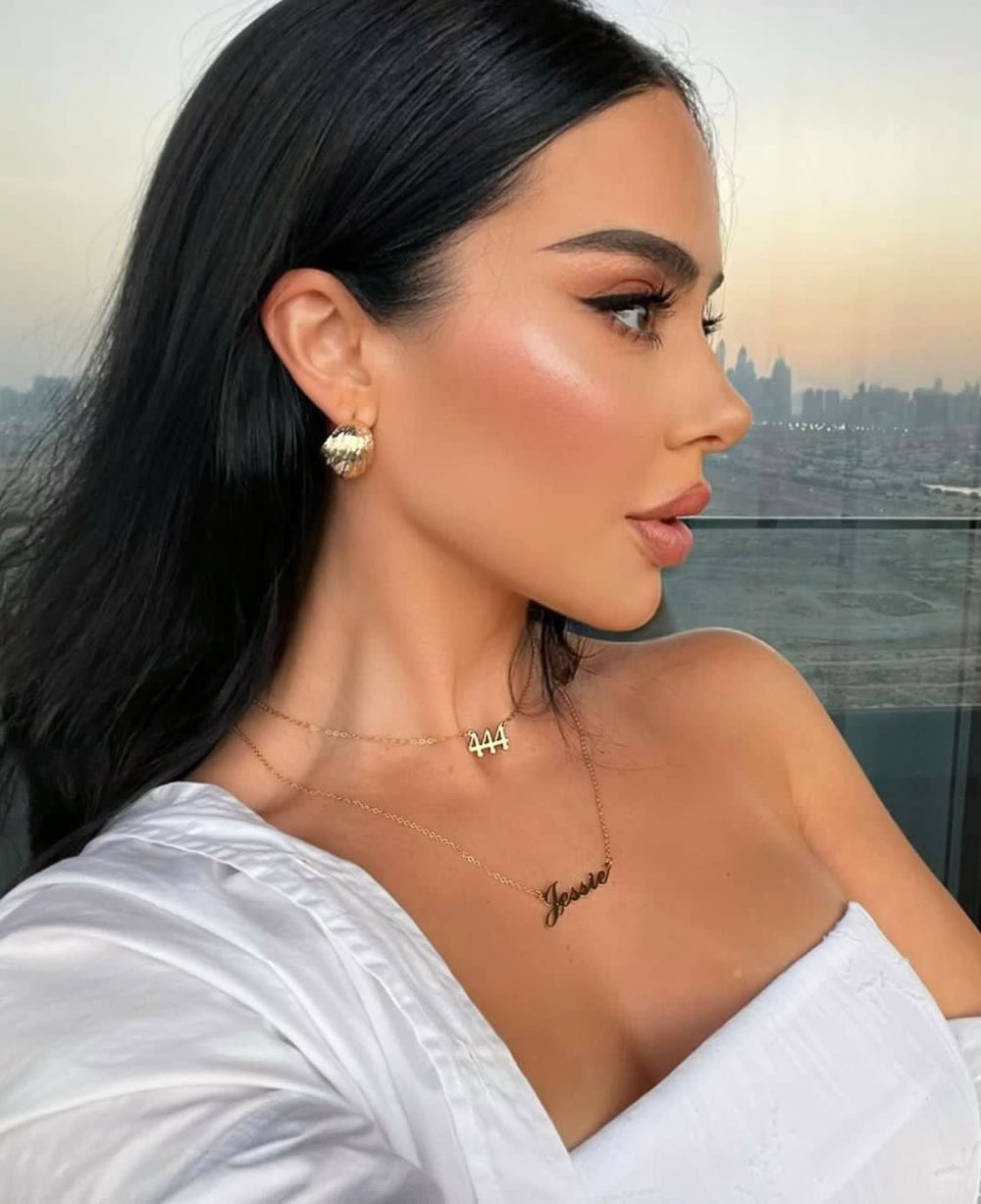 A woman with long dark hair wearing a white dress and gold jewelry, including earrings and a necklace, is shown in profile against a cityscape background.