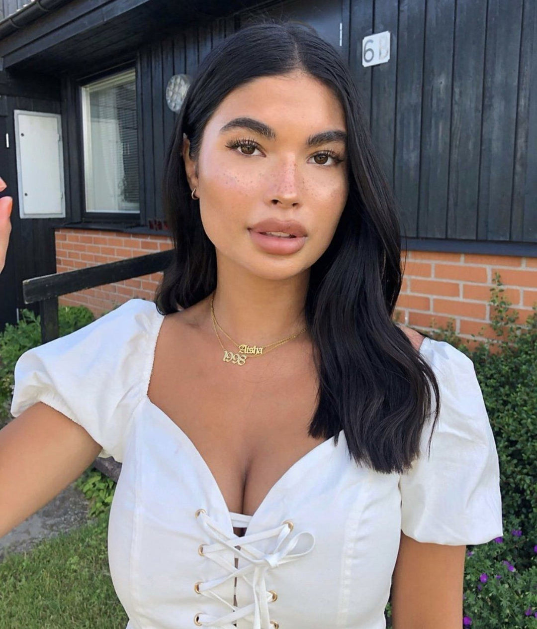 A young woman with long dark hair wearing a white top and posing in front of a brick building.