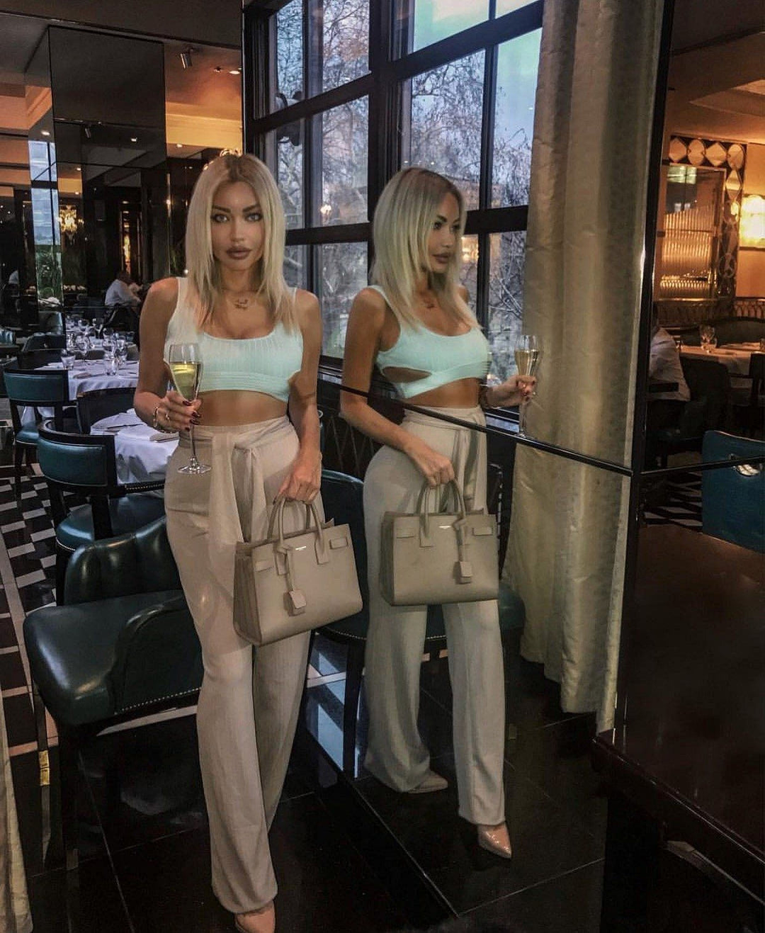 Two women wearing matching crop tops and beige pants are standing in a restaurant, each holding a drink and a handbag.