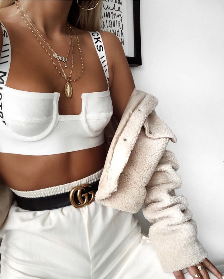 A woman wearing a white crop top, white pants, and a beige jacket, accessorized with a gold belt and multiple necklaces.