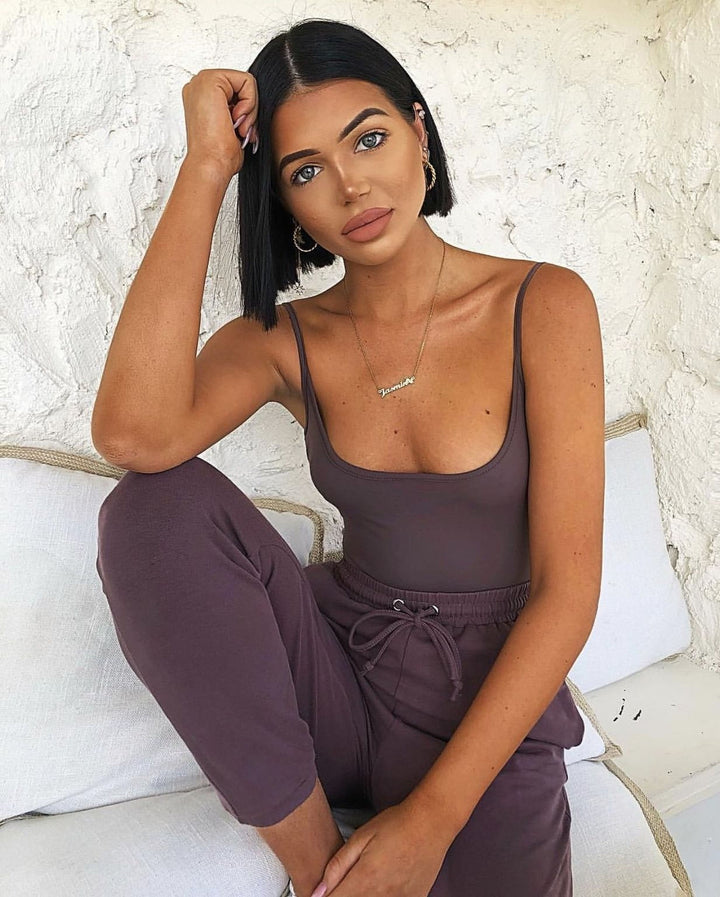 A woman with short dark hair is sitting on a white couch, wearing a purple tank top and matching pants. She has her hand on her head and is looking directly at the camera.