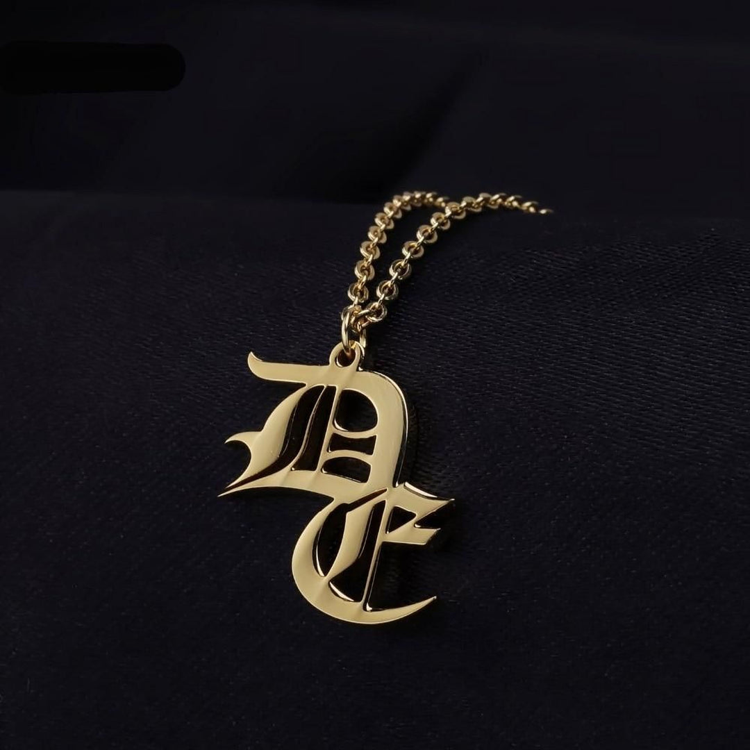 A gold-colored pendant necklace with an ornate, gothic-style letter design hanging against a dark background.