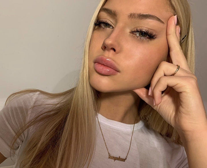 A close-up portrait of a young woman with long blonde hair, wearing a white shirt and a necklace. She is looking directly at the camera with a thoughtful expression.