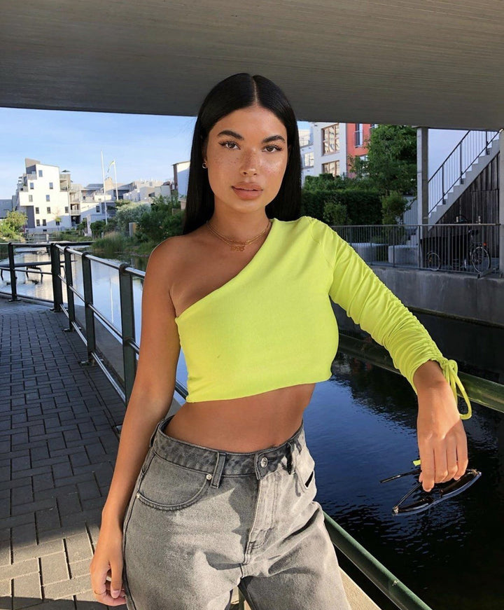 A young woman wearing a bright yellow one-shoulder top and gray jeans stands on a bridge overlooking a body of water, with a cityscape in the background.