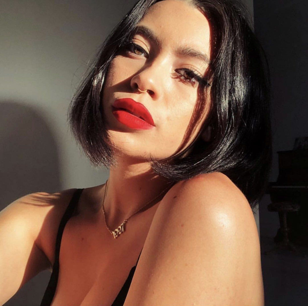 A woman with long dark hair and red lipstick is the main subject of the image. She is wearing a black top and has a pensive expression on her face.