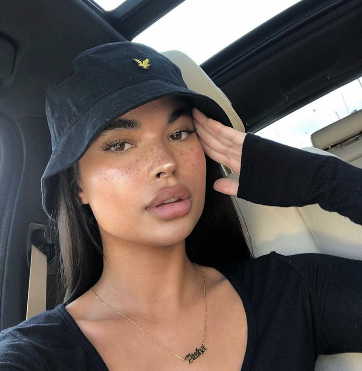 A young woman wearing a black bucket hat and a black top is sitting in a car, with her hand on her face.
