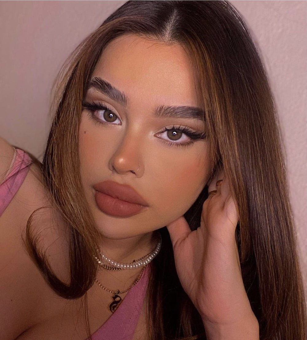 A close-up portrait of a young woman with long, dark hair and striking makeup, wearing a pink top and a pearl necklace.