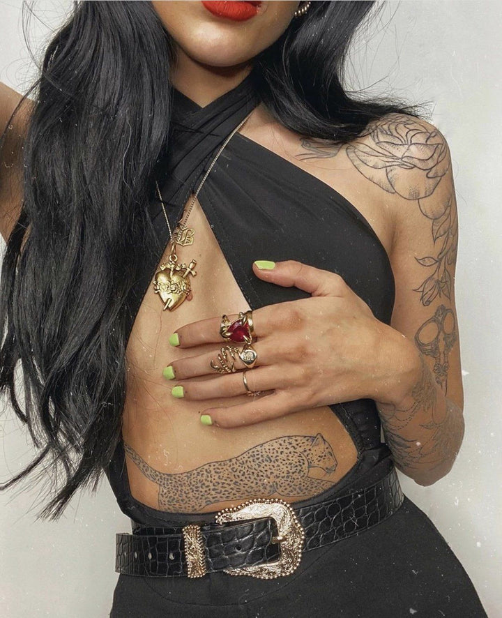A woman with long dark hair wearing a black halter top and a black belt, with tattoos on her arms and a heart-shaped pendant necklace.