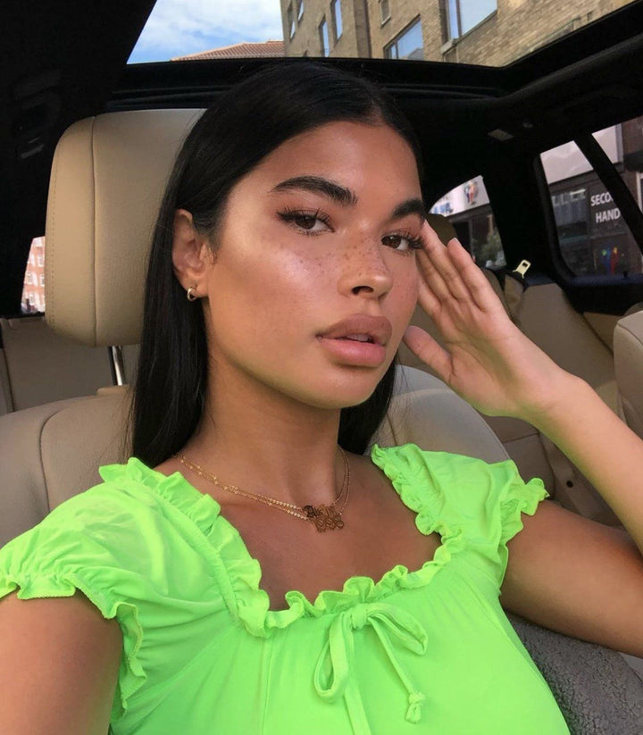 A young woman with dark hair and freckles is sitting in the back seat of a car, wearing a bright green top. She is looking directly at the camera with a serious expression.