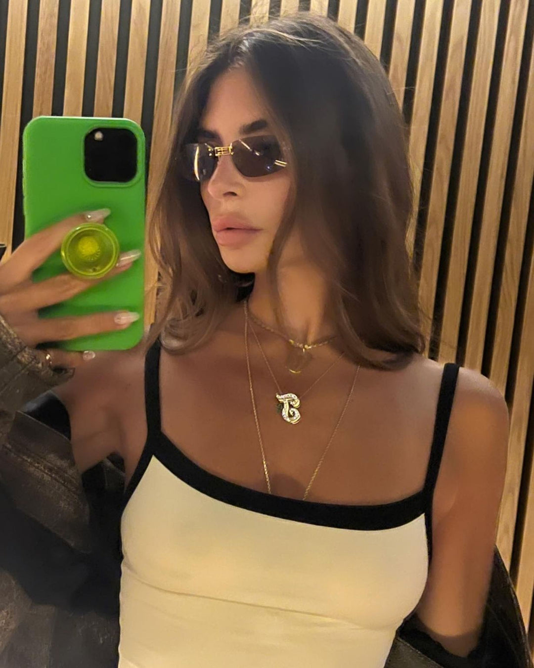 A woman wearing sunglasses and a white tank top is taking a selfie with a green phone case.