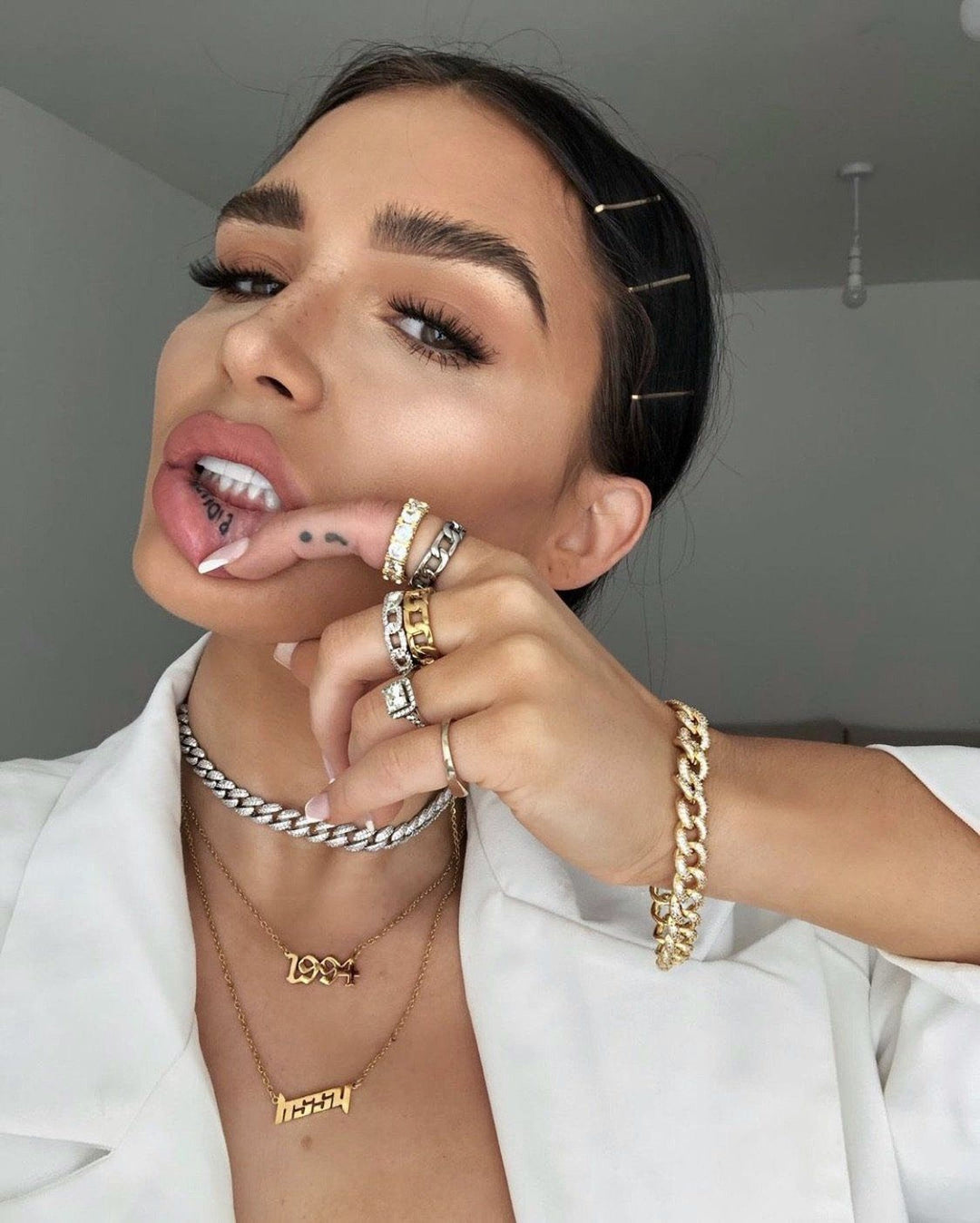 A woman wearing a white jacket and multiple gold necklaces poses with her tongue sticking out, her fingers touching her lips.