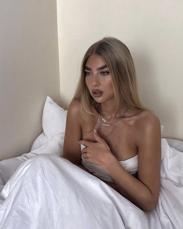A woman with long blonde hair is sitting on a bed, wearing a white top and holding her hands to her chest.