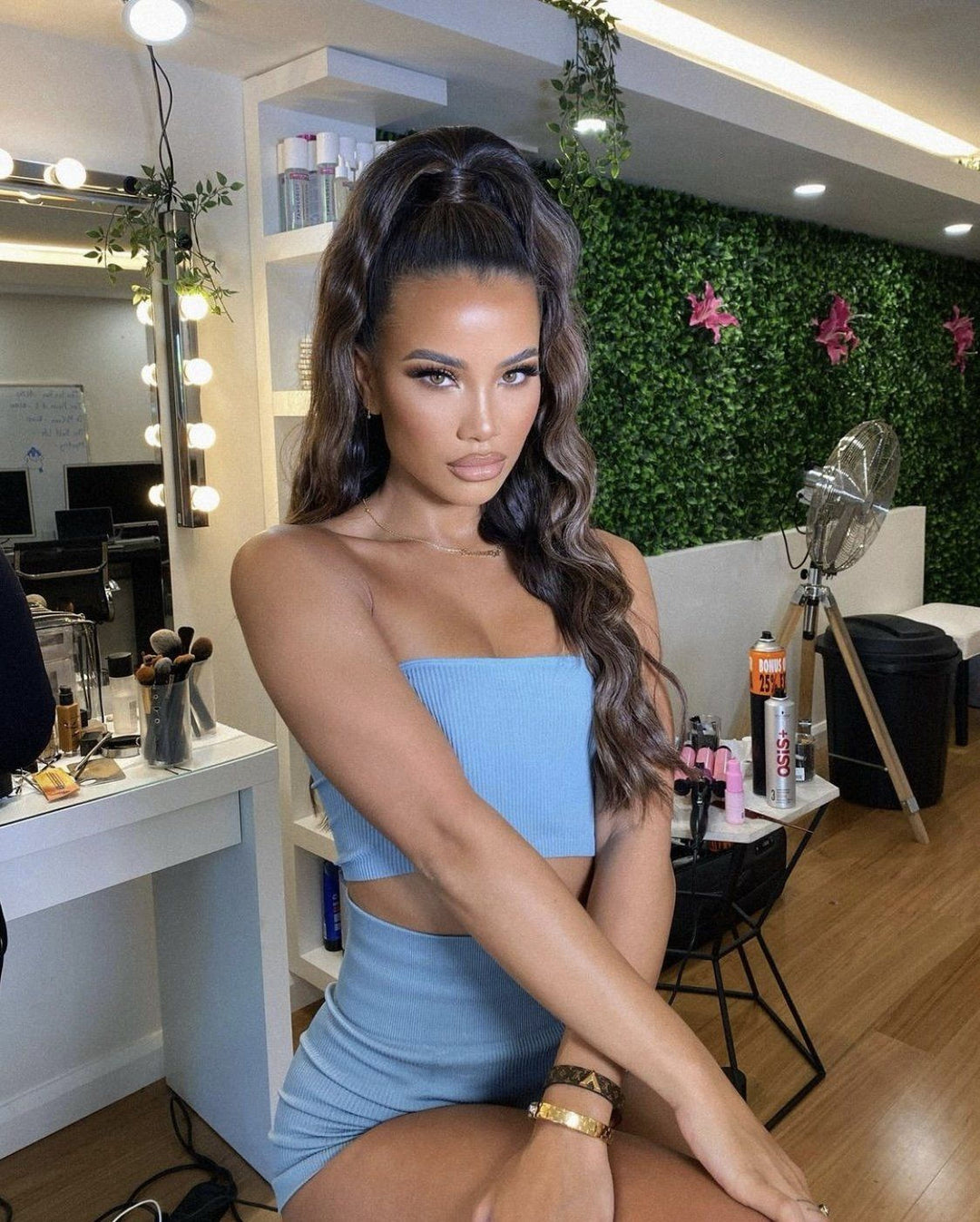 A woman with long dark hair wearing a blue crop top and shorts is posing in a room with a vanity, hair styling tools, and a green wall with pink flowers.