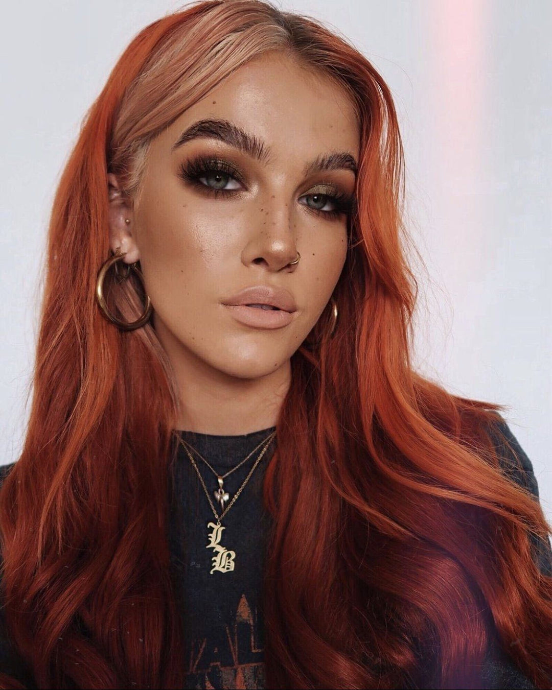 A close-up portrait of a woman with long, reddish-brown hair and striking makeup, including dark eye makeup and bold lipstick. She is wearing large hoop earrings and a necklace with a pendant.