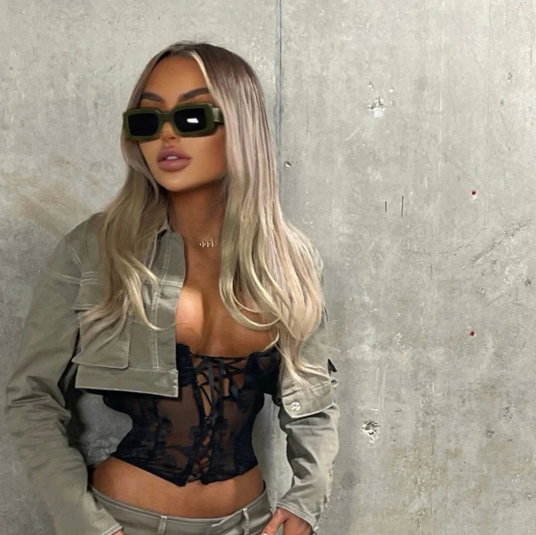 A woman with long blonde hair wearing a black lace-up top and sunglasses stands in front of a concrete wall.
