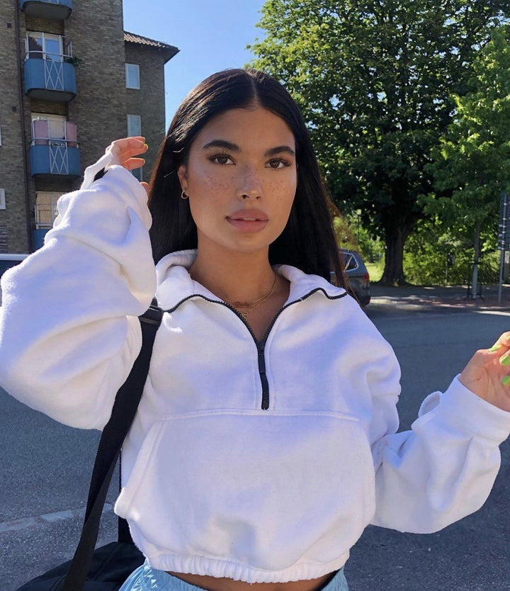 A young woman with long dark hair stands in front of a building, wearing a white hoodie and holding a black bag.