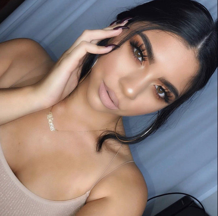 A close-up portrait of a woman with dark hair and makeup, wearing a beige tank top. She is holding her hand to her face, and the background is a blue curtain.