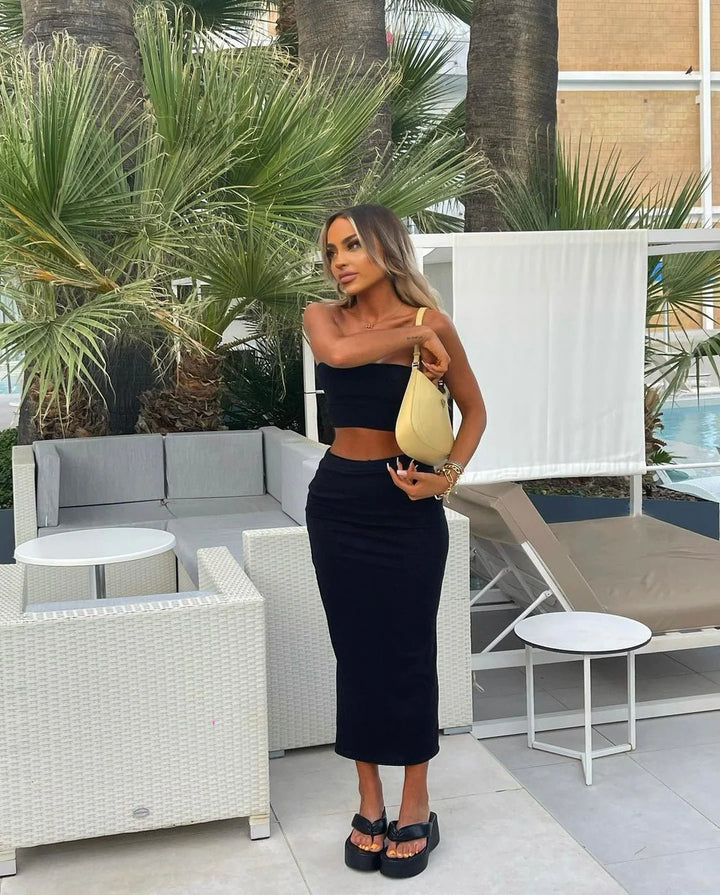 A woman in a black dress and heels stands in an outdoor setting with palm trees and a pool in the background.