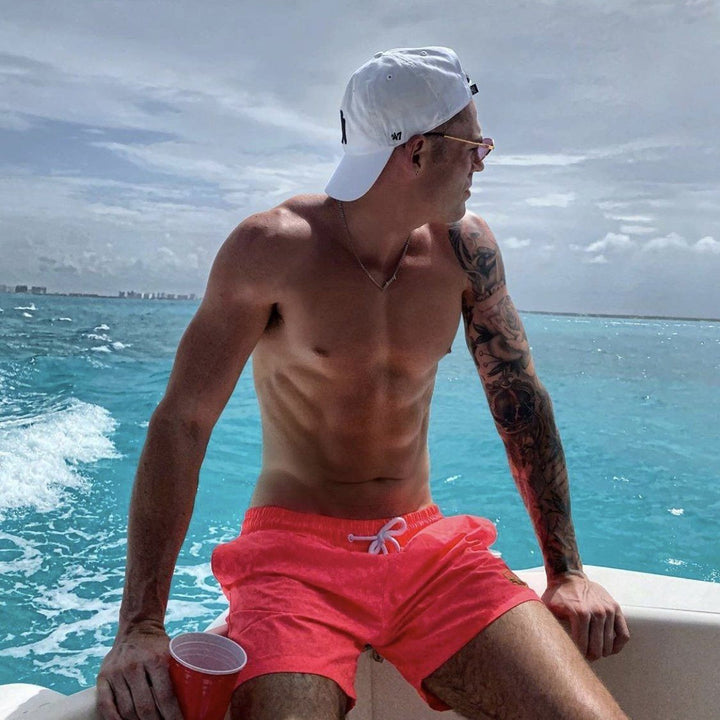 A shirtless man wearing a white cap and sunglasses sits on a boat, holding a red cup, with the ocean and cloudy sky in the background.