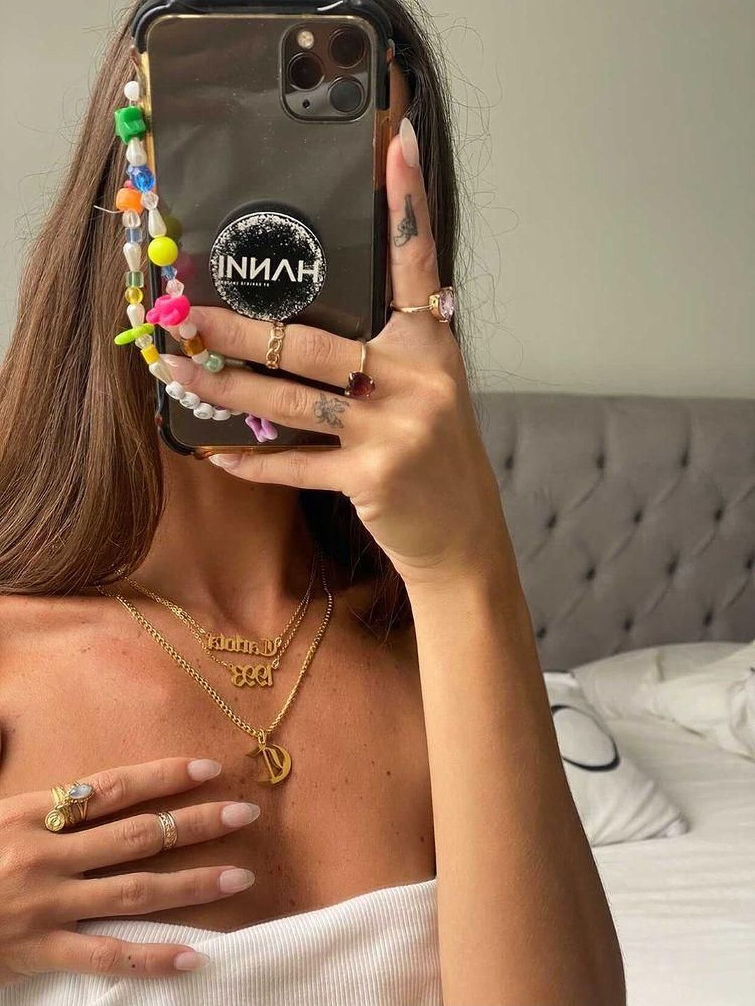 A person is taking a selfie with a phone, wearing multiple rings and necklaces, and holding a colorful beaded charm.