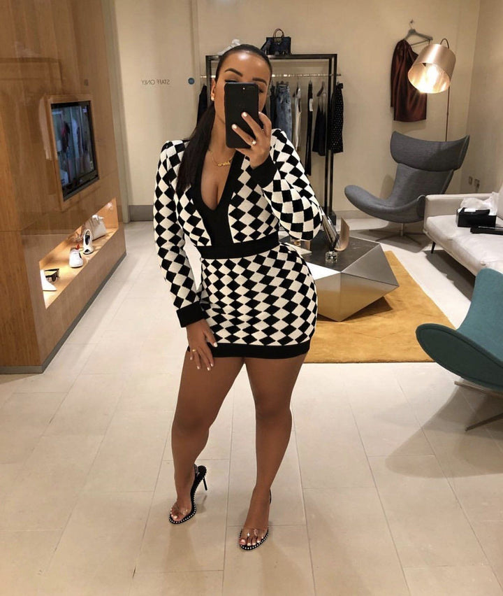 A woman wearing a black and white checkered dress and high heels is taking a selfie in a modern living room with a television, chair, and other furniture.