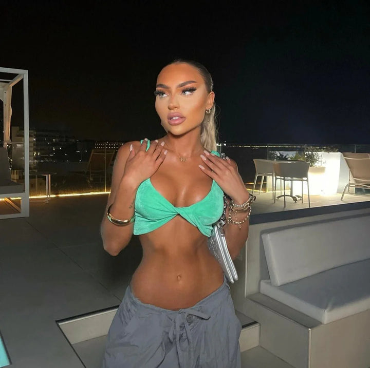 A woman wearing a green top and gray pants stands in a dimly lit outdoor setting, with a city skyline visible in the background.