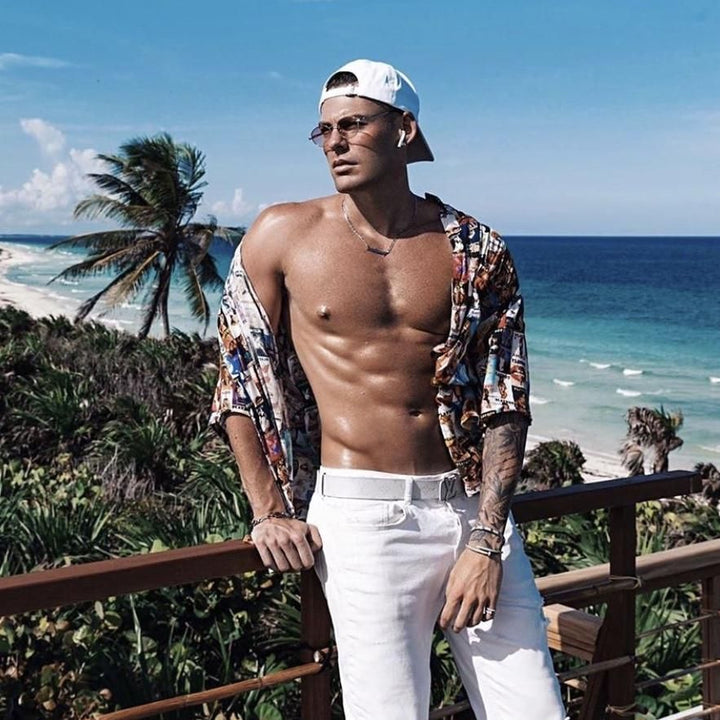 A shirtless man wearing a cap and sunglasses stands on a wooden deck overlooking a tropical beach with palm trees and a clear blue sky.