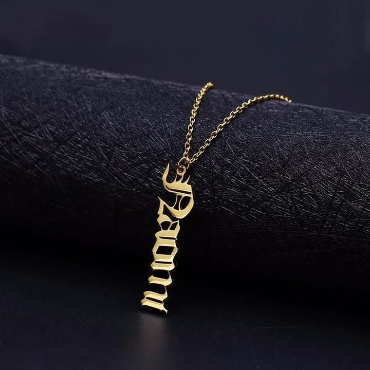 A gold-colored necklace with a pendant in an old-fashioned font, resting on a dark surface.