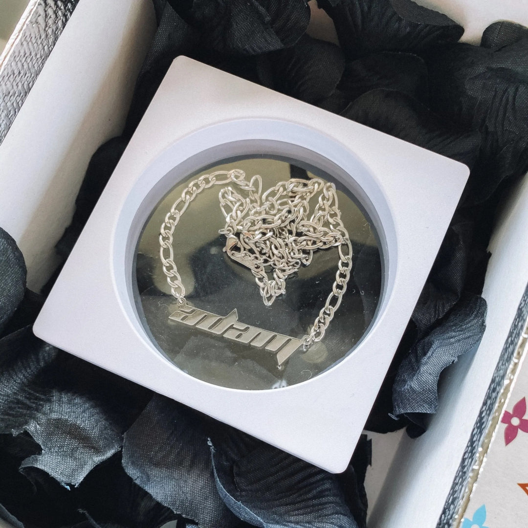A white square box containing a silver chain necklace with a pendant, placed on a bed of black petals.