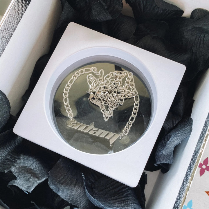 A white square box containing a silver chain necklace with a pendant, placed on a bed of black petals.